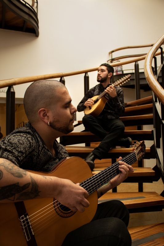 David Valencia toca la guitarra en primer plano mientras Brayan Ruiz toca la bandola sentado en una escalera de caracol